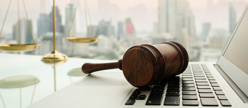 law legal technology concept. Wooden gavel on laptop keyboard with scales of justice in business city background.