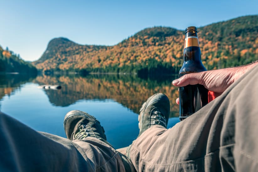DSLR picture POV of a man relaxing near a lake on a beautiful day of autumn season. There is coloured mountains in background and the lake is calm. The man is holding a beer bottle.