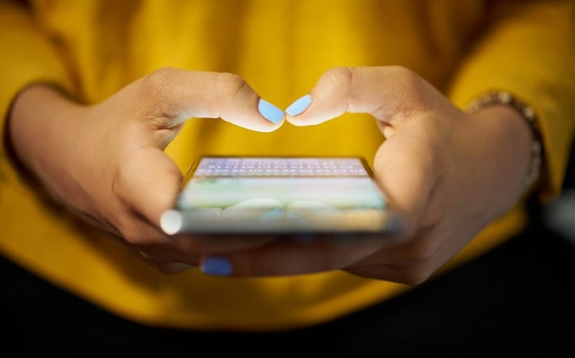 Young woman using cell phone to send text message on social network at night. Closeup of hands with computer laptop in background