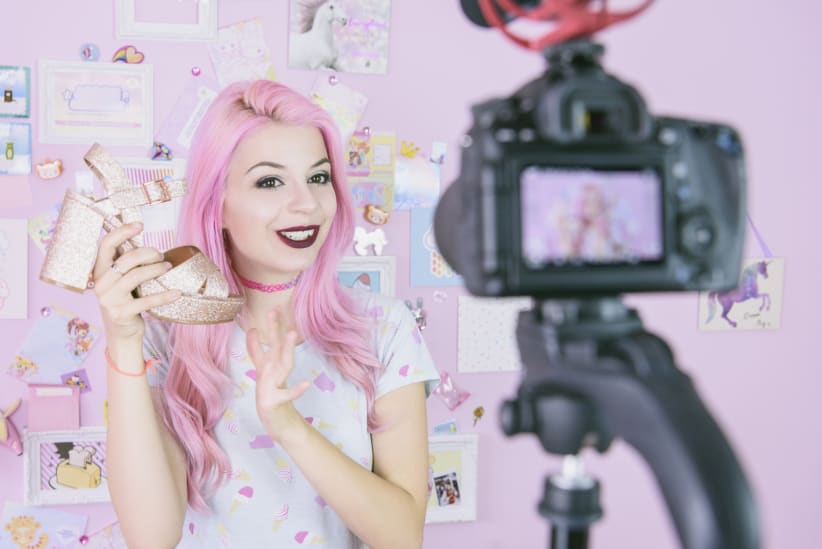 Pink-haired woman holding up a shoe in front of a camera.