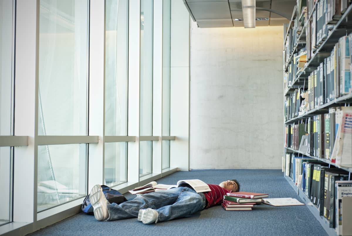 Student laying on the floor of a library, exhausted and surrounded by books