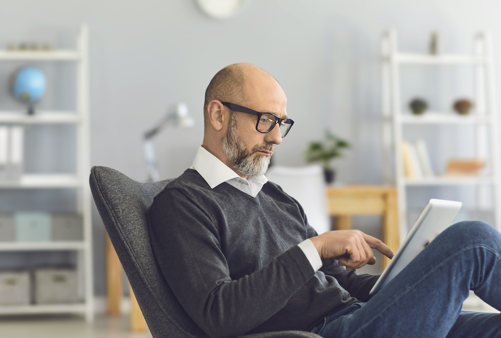 A smart mature man with glasses looks in a tablet in his hands while sitting in a comfortable chair in the living room.